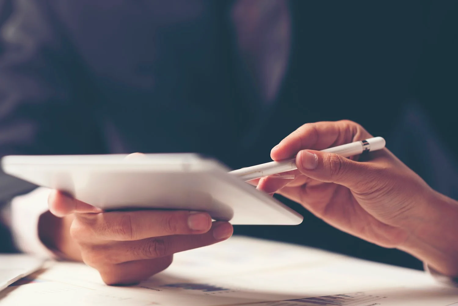 Two hands use a stylus on a tablet, with papers visible beneath it.