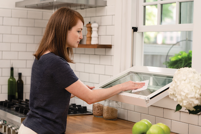 A woman with red hair and a navy shirt cleaning the outside of a single-hung window bottom sash in a kitchen window. 