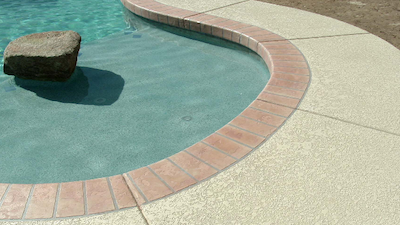 The corner of a rounded swimming pool with red tile around the pool and a beige colored concrete with texture around the pool.