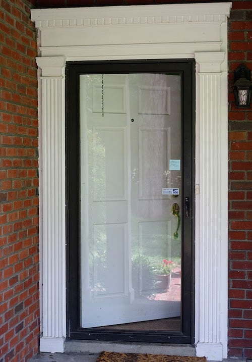 The front of a home with a white door and a storm door in front with a black frame. The door has white moulding around it and red brick on the walls. 