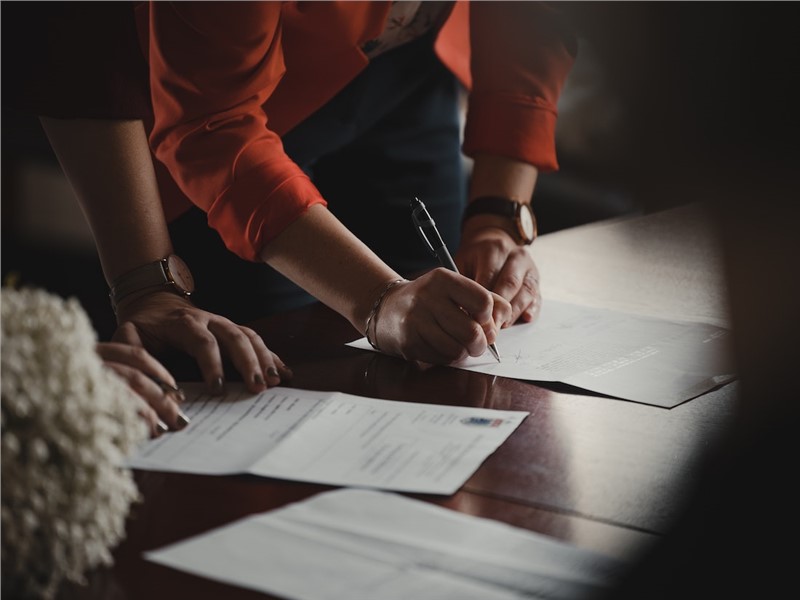 Viewed from over someone's shoulder, the arms of two people leaning against a table looking at papers. The person on the right is wearing an orange long-sleeve shirt and has a pen in their hand up to the paper.