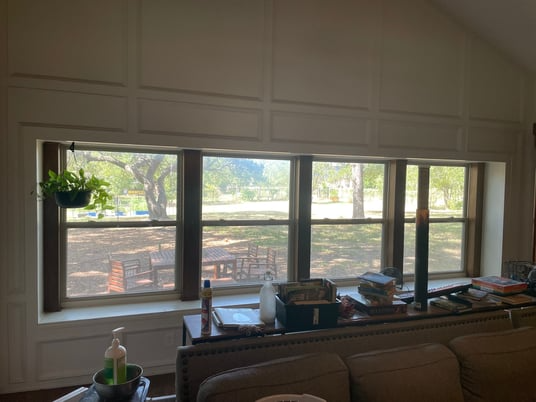 A room features a large window with brown trim. The window looks out onto a backyard with trees, a fence, and patio furniture.