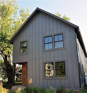 A house with vertical grey siding and wood posts in the front with black windows and a forest background.