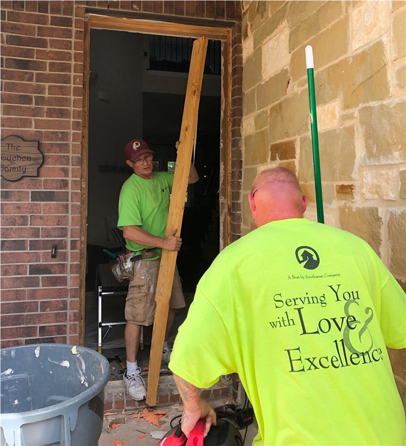 Two men in bright neon yellow shirts in an open doorway replacing the door system. One man is walking out the door with a long wooden two by four and the other man has his back to the camera walking towards the door. There is red brick around the doorway.