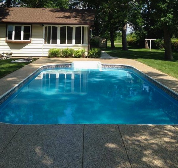 A backyard with a rectangular swimming pool with concrete surrounding. There is a house in the background with green grass and tall trees.