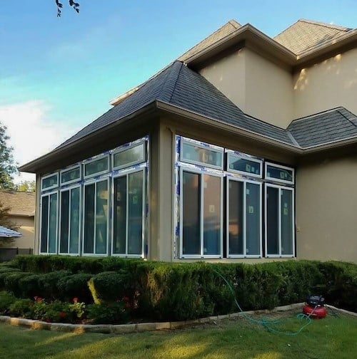 The corner of a house with 8 tall windows that come together on the corner. The house is a tan color with a brown roof. 