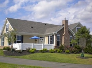 A single-story house with tan siding, dark shutters, a white fence, and a grey roof.