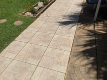 A sidewalk made of tan colored tiles with a geometric pattern surrounded by green grass and some stepping stones leading away.